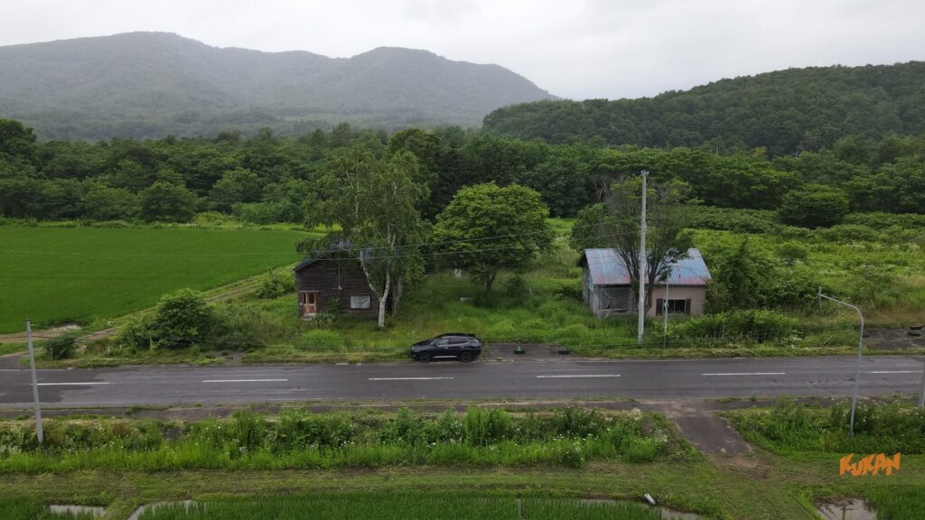 キロロ・ニセコエリアの舗装道路沿い、黒いSUVと2軒の古家付別荘開発用地。霧深い山並み