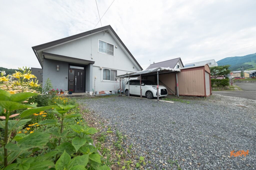 富良野北の峰にある投資用角地戸建て。カーポートと物置のある砂利敷きスペースと玄関外観。