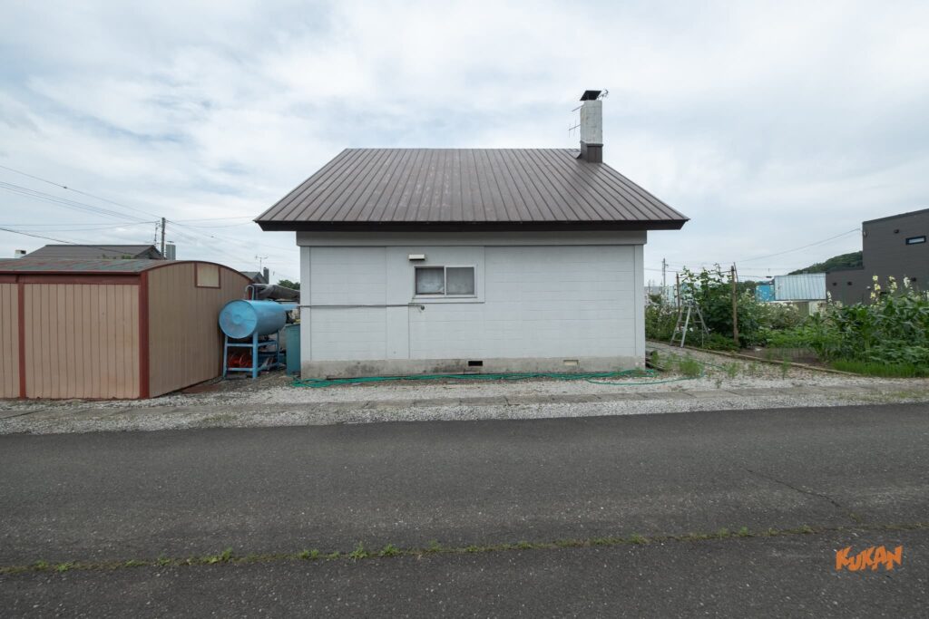 富良野北の峰にある投資用角地戸建ての側面外観。煙突のある屋根と隣接する物置・灯油タンク。