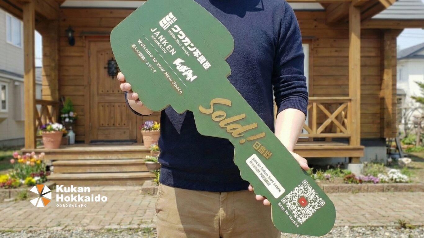 不動産売買の成約を記念した大型鍵型のロゴ入り成約パネルデザイン制作事例。住宅前で「Sold」と書かれたグリーンのオリジナルパネルを持つイメージ。