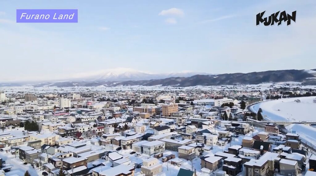 冬の雪に包まれた北海道富良野市の街並みと遠方にそびえる雪山の鳥瞰空撮写真。左上に「Furano Land」のテキスト、右上に「KUKAN」のロゴ。